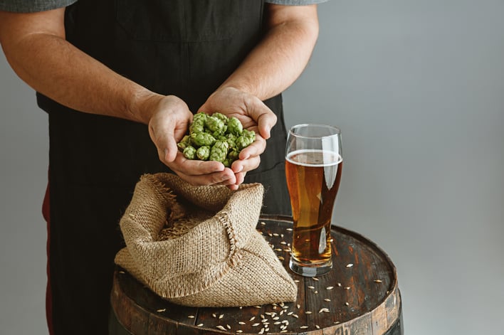 close-up-of-confident-young-man-brewer-with-self-crafted-beer-in-glass-on-wooden-barrel-on-grey-wall